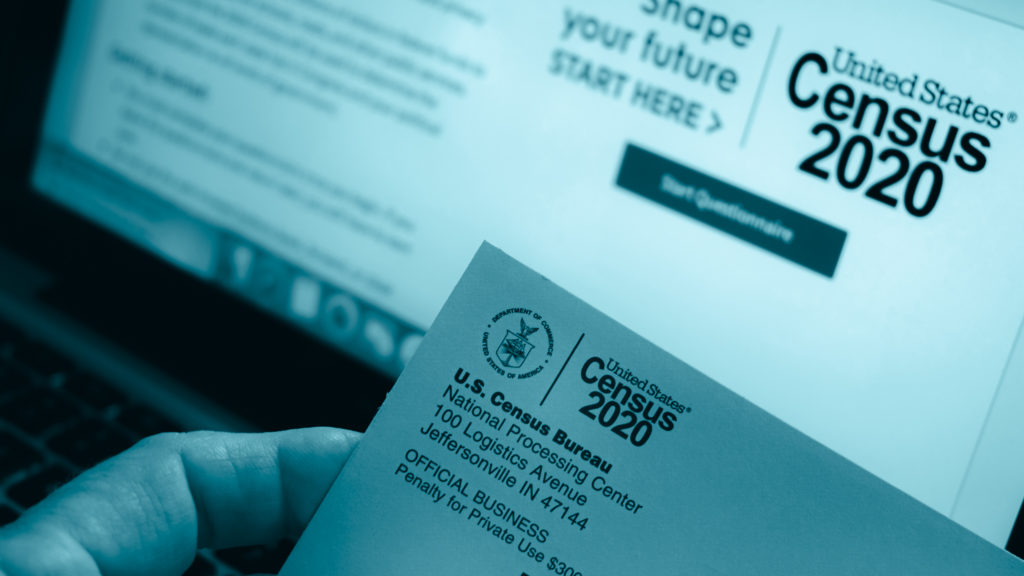 A hand holding a 2020 Census postcard in front of a computer screen on the 2020 Census website.