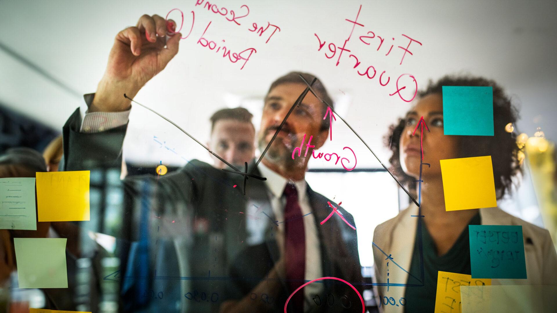 Two people using a clear white board and writing notes